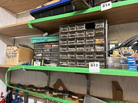 Multi-drawer plastic storage organizer with assorted fasteners on a wooden shelf, alongside boxes, cables, and miscellaneous shop items.