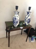 Two tall pottery lamps with blue and white floral patterns placed on a small wooden table with a green leather top. A green bird figurine is also on the table.