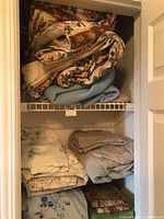 Closet shelves filled with assorted linens including feather duvet, blankets, sheets, pillowcases, and table linens with visible age wear.