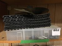 Stack of black interlocking vinyl floor mat squares atop a clear plastic container holding assorted stainless steel clevis pins and bolts.