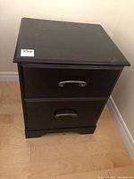 Black night table with two drawers closed, showing front and top surfaces, placed on light wood floor.