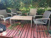 Full patio set showing four metal chairs with woven seats and backs arranged around rectangular metal table with slatted top on red deck with leaves and vine growth.