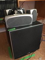 One Polk Audio large black subwoofer with five silver-gray speakers atop it, including one medium and four small speakers, cables present, placed on a wooden floor near shelving.
