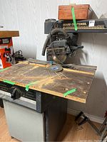 Radial arm saw with attached motor and dust hose, mounted on a worn wooden work surface and metal stand.