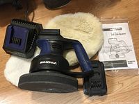 Simoniz cordless buffer/polisher with battery and charger sitting on a white polishing pad on a wooden surface.