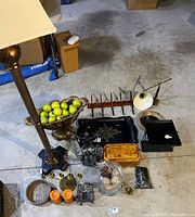 Photo showing a 5ft tall floor lamp with a cream fabric shade standing next to a cluster of home decor items on the floor including a decorative metal bowl with faux green apples, wooden tray, brass candlesticks, orange candles, and various small accents.