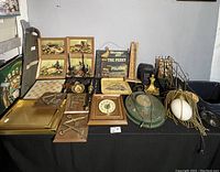 Overview of the entire collection showing paintings, clocks, brassware, metal plaques, and decorative items on a black tablecloth.