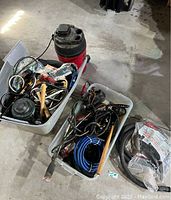 Two bins on concrete floor containing assorted tools, cords, and a wet/dry vacuum cleaner.