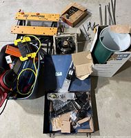 Lot contents overview showing Workmate 200 saw table, aluminum coil in open box, various metal rods, extension cords, and miscellaneous hand tools in bins and on floor.