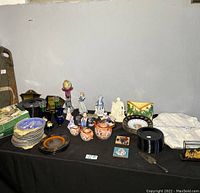 Full view of all items on a black tablecloth, showing plates, figurines, glass vases, ceramic jugs, and additional small decor items arranged together.