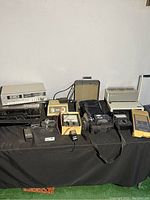 Wide view showing various electronics, camera, typewriter, cassette players and accessories on a black cloth surface.