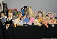 Full view of collection of glass and porcelain dolls arranged on a black cloth surface against a plain wall, showing various sizes and colors of dolls in different costumes.