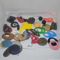 Photo showing 22 pairs of modern earrings laid out including wood, plastic, metal, and shell materials in multiple bright and natural colors and various shapes like ovals, circles, hearts, and geometric forms.
