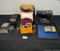 Full shot showing all items on black table including Toshiba laptop, Casio package, camera equipment in box, radios, GPS, and leather camera case.