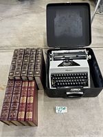 Full view of the Olympia typewriter in its black carrying case next to four large bound books including Eaton Press Americana encyclopedia 2 volumes and two other leather-bound books.