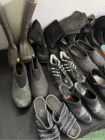 Photo of various boots, clogs, athletic shoes, and slip-ons in black, brown, gray, and beige on black floor.