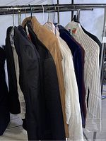 Multiple women's blazers and button up shirts hanging on rack showing variety of styles and colors including black blazer, tan fringe jacket and striped button ups