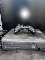Black Xbox 360 console with Xbox 360 controller on top, front view showing power button and disc tray