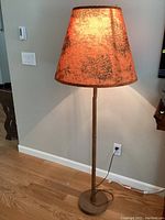 Full view of floor lamp with orange silk shade illuminated against beige wall and hardwood floor