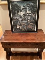 Framed painting resting on top of carved wooden cabinet, showing overall look and size.