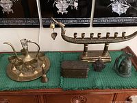 Wide view of lot including brass menorah, Moroccan tea set with tray and cups, decorative metal box, ornate flask, and dragon bell all arranged on green cloth on wooden surface.