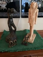 View of all four items with pharaoh statue, nude statue showing chipping, and two small metal figurines on a green cloth.