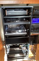 Front view of black cabinet with glass doors showing stereo equipment inside including Nakamichi cassette player, Sony Blu-Ray player, turntable, and other components.