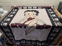 Full view of Betty Boop throw blanket spread on table showing central Betty Boop image and surrounding border with smaller face images and fringe edges.