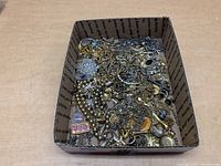 Box filled with mixed jewelry including chains, bracelets, pendants, and earrings in silver and gold tones.