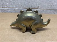 Front and side view of the brass dinosaur coin bank showing the stegosaurus shape, smooth polished surface, and details of the legs, tail, and head.