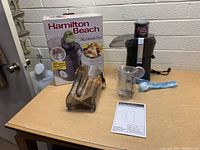 Photo showing the boxed Hamilton Beach Big Mouth Pro Juicer along with the main juicer unit, pulp collector, juice pitcher, instruction manual, and cleaning brush all displayed on a wooden table.