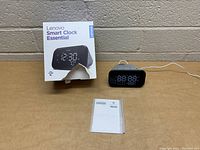 Photo of Lenovo Smart Clock Essential with original box and user manual, clock plugged in and upright on a flat surface.