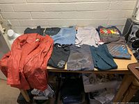 Overview of the lot showing the red leather jacket, various t-shirts, denim jeans, and knitwear on a table in a room with a white brick wall.