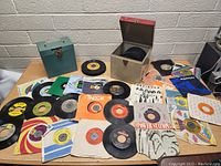 Photo showing spread of approximately 30 vintage 7-inch vinyl records in paper sleeves, some with colorful labels and multiple artists visible, including Foreigner. Two carrying cases (one metallic and one teal green) are visible.