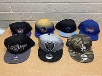 Overview of 6 assorted snapback hats featuring NBA and NFL teams arranged on table, showing front logos and colors.