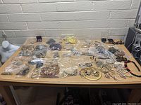 Photo showing a wooden table with many small plastic bags containing assorted jewelry pieces including necklaces, bracelets, and earrings. Some loose necklaces and bracelets displayed at front.