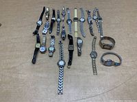 Overview image showing 15 wrist watches assorted on cardboard surface in various styles and materials including leather, metal link, and expandable bands.