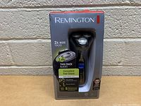 Front view of boxed Remington rotary shaver showing branding, product window, and features like twin track blades and consistent fast shave.
