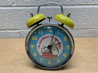 Front view of the vintage 1958 Snoopy Peanuts alarm clock showing the blue face, yellow numbers, and Snoopy illustration with the phrase 'I'M ALLERGIC TO MORNING'. Yellow twin bells on top against a white wall background.