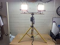 Front view of the two illuminated work lights mounted on the yellow tripod stand placed on a wooden table.