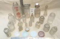 Front view of 18 antique and vintage small glass bottles and jars of varying sizes, with a measuring ruler for scale showing small to medium bottles. Clear glass with some vintage markings and closures.