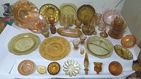 Photo of the full array of amber, peach, and pink Carnival Depression glassware arranged on a table showing plates, bowls, cups, and decorative pieces.