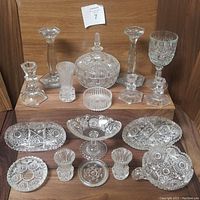 17 pieces of cut lead crystal starburst formal dinnerware including candlesticks, covered jar, goblet, footed bowl, plates, vases, and small dishes, displayed on wooden shelves