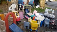 Photo shows an outdoor assortment of multiple children's toys and play equipment including Little Tikes cozy coupe ride-on car, several plastic slides, plastic picnic table with chairs, toy kitchen, baby bath with bed, and a toy shopping cart. Items are heavily used and show dirt.