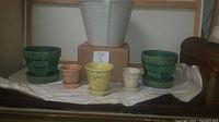 Front view of six vintage pottery planters arranged on a cloth: three green McCoy planters with ribbed texture and three smaller with beige, yellow, and cream colors. Large white planter in background on a box.