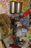 View of MEC 600 Jr. shell reloader with two powder/shot hoppers, assembled with red handle lever, alongside boxes of yellow shotgun shell cases and packets of reloading supplies.