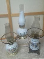 Photo showing three milk glass lamps with floral painted globes; one oil lamp with frosted chimney and two electric lamps with clear chimneys and brass fittings.