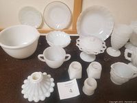 White milk glass dishes and serveware arranged on brown cloth surface, includes hobnail pattern vase, pedestal cake stand, small plates and handled cups.