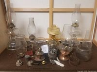 Wide shot showing full lot of glass lamp globes, metal bases, burner parts, and accessories arranged on wooden surface.