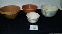 Four pottery bowls displayed with a black cloth background. Two cream-colored bowls with horizontal blue and pink stripes, one small and one large, positioned front center and right respectively; two brown textured bowls behind them, one with stepped ridges and the other with basket weave pattern.